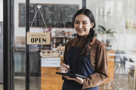 Portrait of a beautiful Asian woman in an apron standing using a tablet in a cafe, she owns a coffee shop, the concept of a food and beverage business. Store management by a business woman.の写真素材
