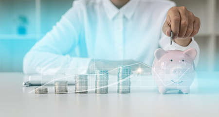 A person puts a coin in a pig piggy bank, a large number of coins are placed on the table from low to high, chart graphics showing the financial situation. concept of personal finance managementの写真素材