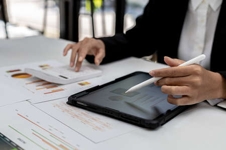 A woman looking at a tablet at a screen showing company financial status chart and presses a calculator to check, a finance worker is checking documents. Concept of company financial management.の写真素材