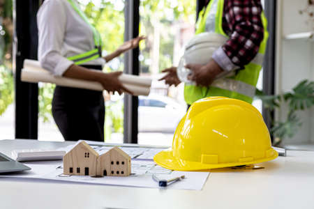 Helmets and miniature house models sit on the desks of engineers and architects designing interiors of houses and buildings with two people standing behind them talking. Interior design ideas.の写真素材