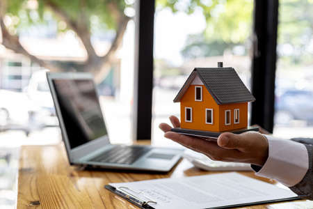 Salesman holding an orange model of a small house, a housing project salesman is drafting a sales contract for a customer who reserves a house in a project he maintains. Real estate trading concept.の写真素材