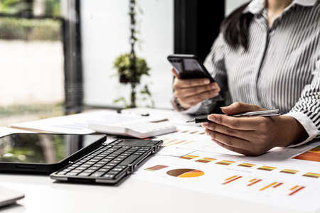 Person holding a mobile phone and pointing a pen at the document, the finance staff is reviewing the information of the company's financial documents to conclude a meeting with the management.の写真素材
