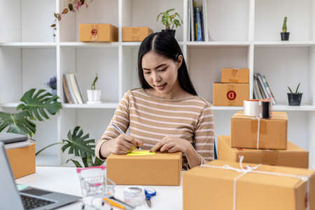 Asian woman writing customer's shipping information on parcel box, she owns an online store, she...