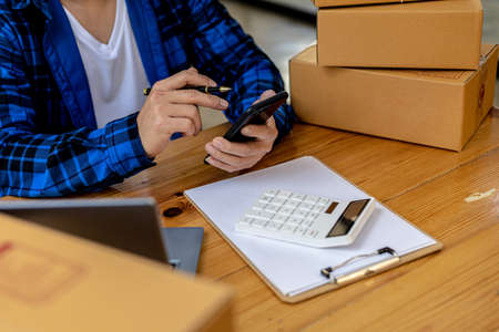 A male shopkeeper answering customer chats via mobile phones, He owns an online store, He packs and ships through a private transport company. Online selling and online shopping concepts.の写真素材