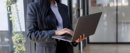 A businesswoman with a laptop standing by the window, she owns a startup company, a young and energetic businesswoman. Concept of business woman, female leader.の写真素材