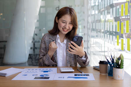 Businesswoman showing joy and holding a mobile phone. She checked the sales on her mobile phone, she was glad the sales were increasing so she showed a happy expression. Sales management concept.の写真素材