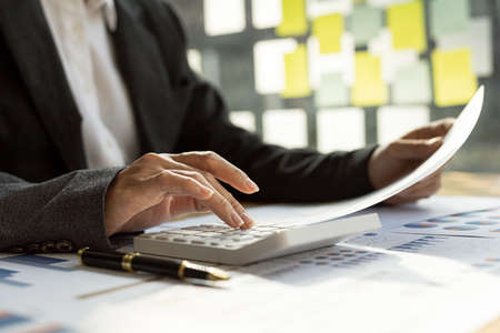 Businessman using a calculator to calculate numbers on a company's financial documents, he is...