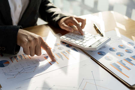Businessman using a calculator to calculate numbers on a company's financial documents, he is...