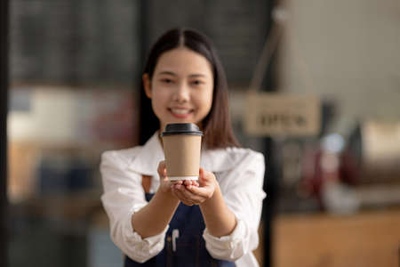 A female receptionist in a cafÃ© holds a paper cup of hot coffee in front of her, she is a cafÃ© employee who serves and serves customers who come to use the service in the shop.の写真素材
