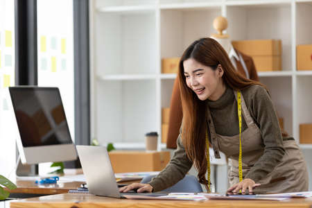 A beautiful Asian woman who owns a clothing brand, she is designing clothes on a laptop, she designs all of her own brands and sells them online and has a storefront. Clothing design ideas.の写真素材