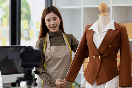 A beautiful Asian woman who owns a clothing brand, she is live chatting with customers and presenting a new collection of clothes, she designs all the clothing of the brand herself. Clothing design ideas.の写真素材