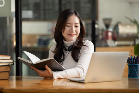 Beautiful Asian woman holding a book and looking at the laptop, she is studying online. The concept of online learning due to the COVID-19 outbreak to prevent an outbreak in the classroom.の写真素材