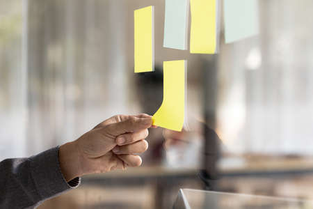 Note paper pasted on glass in meeting room, businessman's hand pointing and pasting note paper to write messages or meeting topics. The idea of writing a notepad to memorize.の写真素材