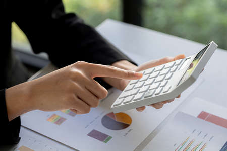 Businessman using a calculator to calculate numbers on a company's financial documents, he is...