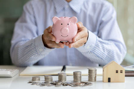 A person is holding a pink pig piggy bank, piles of coins on the table and a house model on it,...