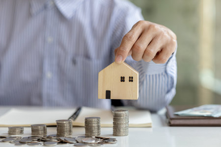 Person holding a small wooden house model, stacks of coins on the table, saving money to spend and managing to buy a future home. Money saving ideas to buy a house or residence.の写真素材
