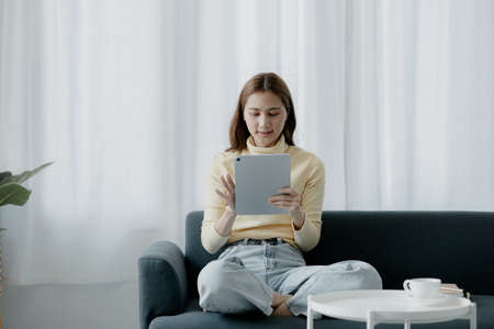 A woman plays a tablet on the sofa in her living room at home, she is resting on weekends after a...