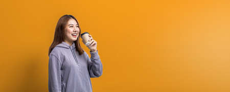 Portrait of beautiful Asian woman holding a paper coffee cup on isolated yellow background, portrait...