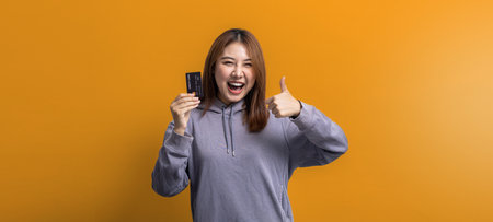 Portrait of Asian woman holding a credit card, credit card spending concept, online payment and general store with credit card, isolated over yellow background, copy spaceの写真素材