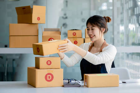 A beautiful Asian business owner opens an online store, she is checking orders from customers, sending goods through a courier company, concept of a woman opening an online business.の写真素材