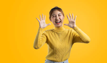 Portrait of a beautiful Asian woman in a yellow shirt standing with her hands up in joy. portrait concept used for advertisement and signage, isolated over the blank background, copy space.の写真素材