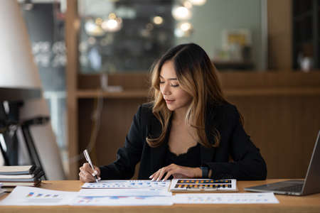 A beautiful Asian businesswoman sitting in her private office, she is checking company financial documents, she is a female executive of a startup company. Concept of financial management.の写真素材