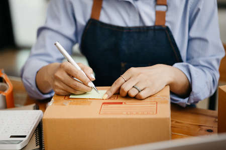 Woman who runs an e-commerce business is writing a list of customers on paper before shipping to...