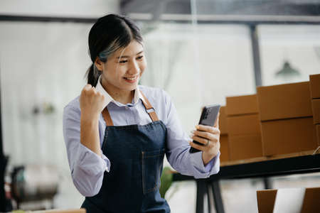 Woman who runs an online store with a happy face is holding her phone to check sales, she sells...