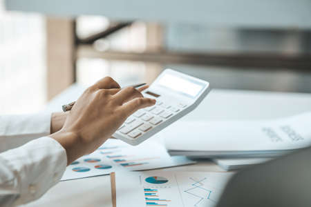 Businessman using a calculator to calculate numbers on a company's financial documents, he is...