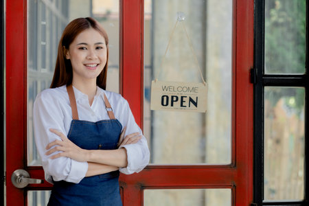 Asian woman in an apron stands holding a restaurant's menu in front of the door with a sign opening a shop, an opening of a small restaurant, a woman running a business. Operating a small restaurant.の写真素材