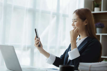A woman raising her hand in a happy gesture and looks at her phone, an Asian businesswoman looking at monthly sales data on her smartphone and looks happy. Sales management concept.の写真素材