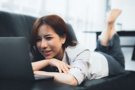 Asian woman lying on the sofa playing laptop on the sofa, she is taking a weekend break after a hard day's work. Holiday relaxation ideas.の写真素材
