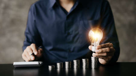 Businessman holds a glowing light bulb on top of the highest pile of coins, Placing coins in a row from low to high is comparable to saving money to grow more. Money saving ideas for investing in fundの写真素材