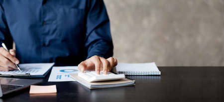 Businessman is using a calculator to calculate company financial figures from earnings papers, a businessman sitting in his office where the company financial chart is placed. Banner with copy space.の写真素材