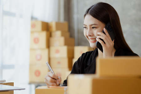 Asian woman is talking on the phone with a customer to confirm an order, she owns an online store, she packs and ships through a private transport company. Online selling and online shopping concepts.の写真素材