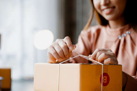 An Asian woman tying a parcel to a customer's box, she owns an online store, she packs and ships through a private transport company. Online selling and online shopping concepts.の写真素材