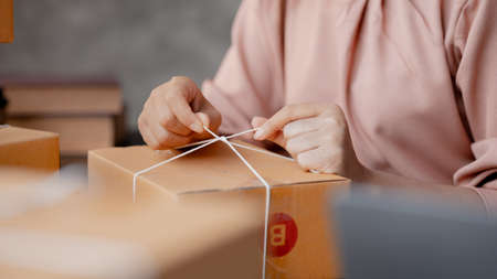 An Asian woman tying a parcel to a customer's box, she owns an online store, she packs and ships through a private transport company. Online selling and online shopping concepts.の写真素材