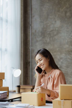 Asian woman is talking on the phone with a customer to confirm an order, she owns an online store,...
