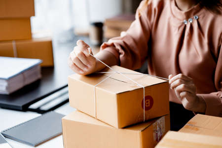 An Asian woman tying a parcel to a customer's box, she owns an online store, she packs and ships through a private transport company. Online selling and online shopping concepts.の写真素材