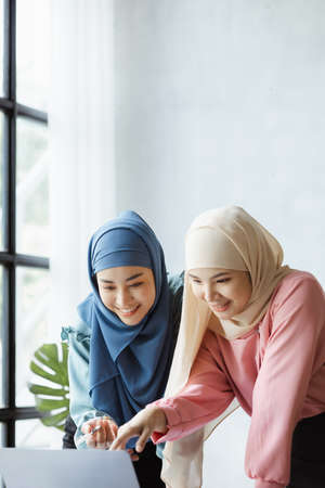 Two hijab-clad Asian women are brainstorming together in a conference room for a startup, run by a...