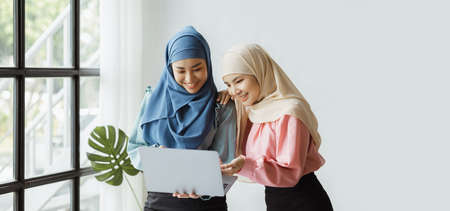 Two hijab-clad Asian women are brainstorming together in a conference room for a startup, run by a...