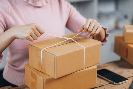 An Asian woman tying a parcel to a customer's box, she owns an online store, she packs and ships...