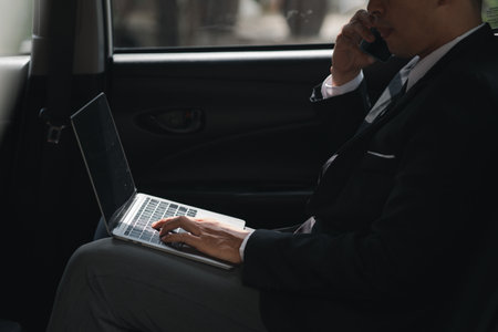 An Asian man in a formal suit is in a car, he is talking on the phone and using a laptop, he sits...