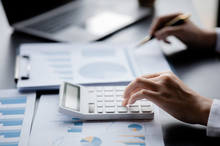 Businessman is using a calculator to calculate company financial figures from earnings papers, a businessman sitting in his office where the company financial chart is placed.の写真素材