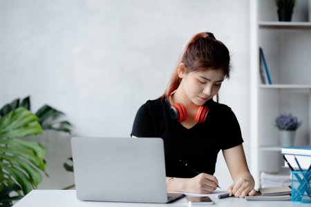 Asian teenage woman sitting in white office with laptop, she is a student studying online with laptop at home, university student studying online, online web education concept.の写真素材
