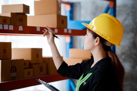 Asian female worker wearing safety clothing is in a warehouse, she is checking the goods in the...