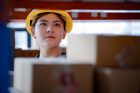 Asian female worker wearing safety clothing is in a warehouse, she is checking the goods in the warehouse before shipping. The concept of working in the warehouse and working in the warehouse is safe.の写真素材