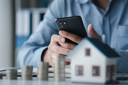 Person holding phone with house model and stack of coins calculating and recording income and expenses for financial planning, financial planning ideas to buy a house and real estate.の写真素材
