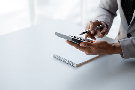 Businessman is using a calculator to calculate company financial figures from earnings papers, a businessman sitting in his office where the company financial chart is placed.の写真素材