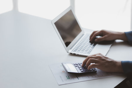 Businessman using a calculator to calculate numbers on a company's financial documents, he is...
